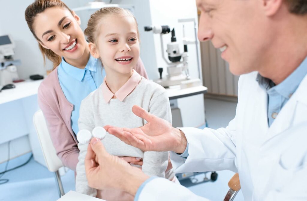 An optometrist holds a contact lens case while explaining myopia control options to a young patient and their mother during a routine eye exam.